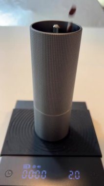 Kahve çekirdekleri modern metal kahve öğütücüsüne dökülüyor. Beyaz bir masanın üzerindeki tüm detayları gösteriyor. Kahve öğütücüsü, kahve yapmak için kullanılan tahıl miktarını tartıyor.