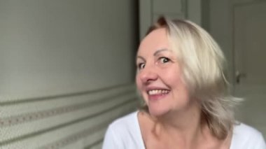 Sarışın kadın başka tarafa bakıyor ve kafasını çerçeveye çeviriyor. Gülümsüyor ve arka planda beyaz bir tişört giyiyor. Hiç makyaj yapmıyor. 35-45 yaşlarında beyaz dişleri var.