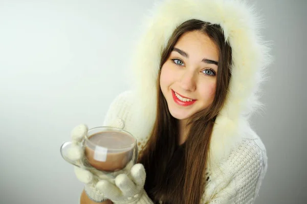 To advertise New Year warming drinks tea and coffee Beautiful young girl drinks from a glass cup of coffee or cocoa White coat and white gloves Snow Maiden with long hair put on a hood Place for text