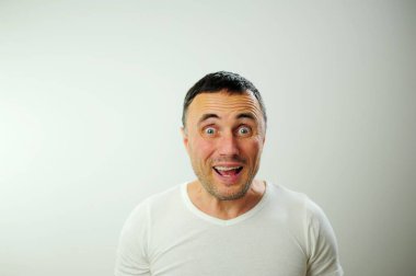 Kırışık alın, dil, farklı neşeli pozitif duygular yetişkin adamın siyah saçı komik mavi gözler beyaz tişört gri arka plan kıllı erkek elleri eğlenceli tatil seyahati kazanır