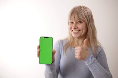 45-50 yaşlarında, mavi elbiseli, sarı saçlı bir yetişkin kadın elinde cep telefonu, yeşil ekran krom anahtarıyla duruyor.