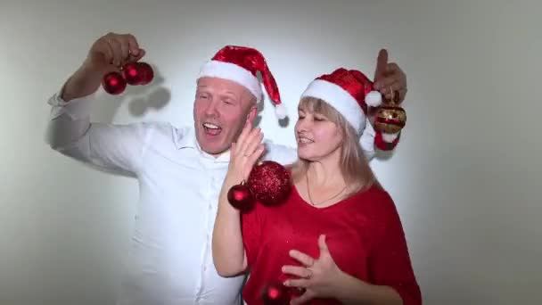 パーティー招待新年とクリスマスの男と女大人はおもちゃで彼らの手で踊る彼らは彼らの頭の上にクリスマスの帽子と赤いボールを持っています — ストック動画