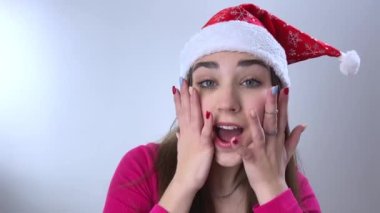Noel Baba şapkalı, Noel Baba şapkalı kırmızı şapkalı genç kız. Gülümsüyor, elleriyle yüzünü tutuyor. Sonra gülüyor, şapkasına dokunuyor.