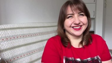 Tombul bir kadın, kıvrımlı ve iştah açıcı, kameranın önünde oturuyor ve gülümsüyor, kırmızı ruju, kırmızı ceketi ve kahverengi saçı var.