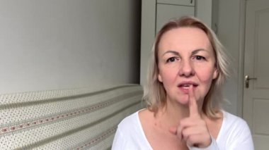 Beyaz ceketli, sarışın, orta yaşlı, yetişkin bir kadın parmağını sallayarak bir şey söyleme. Kapa çeneni kadın, parmağını dudaklarına koy.