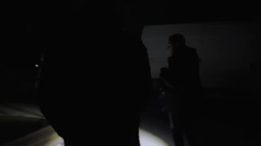 Karanlık hangarda üç kişi yürüyor yere bir el feneri tutuyorlar iki adam ve bir kadın silueti karanlık tuğla tünel ya da kanalizasyon koridorunda feneri olan bir adam. 