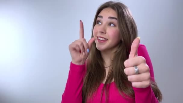 一个有着一头棕色长发的可爱女孩指着一个空的地方发短信 然后挥动她的手指给这个产品提建议 然后转身对着摄像机 把一个大拇指举到课堂上 — 图库视频影像