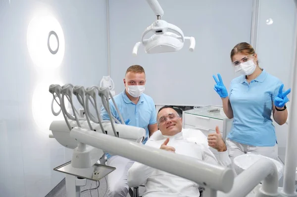 Three joyful and contented patient, doctor, dental assistant thumb up successful dental treatment remove tartar in dental office masks and gloves in blue color transparent glasses in clean stomatology