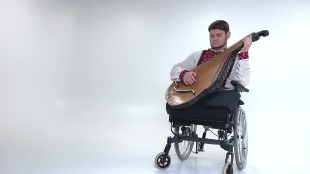 身披科巴扎乐器 体弱多病 英俊的年轻人坐在轮椅上 没有腿 坐在白色的背景上 战争老兵身穿乌克兰绣花衬衫 民族符号为Vyshyvanka — 图库视频影像