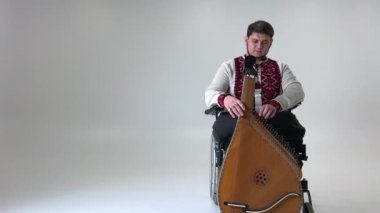 Elinde kobza müzik aleti olan yakışıklı genç adam tekerlekli sandalyede beyaz arka planda oturuyor bacakları olmayan savaş gazisi Ukrayna nakışlı gömlek giymiş vyshyvanka 'nın ulusal sembolleri.