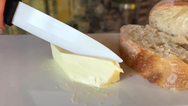 白いナイフの手で白い磁器板の上にバターの一片をカット黄色のバターのいくつかのスライスは まな板上のナイフで大きな部分からカット いくつかのライ麦パン — ストック動画