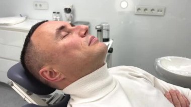 Modern dişçi ofisi ve tıp kürsüsü. Diş hekimliği, orta yaşlı, rahat ve yakışıklı bir adam ağız boşluğu tedavisi için bir sandalyeye ve beyaz ekipmana oturuyor. İç konsept