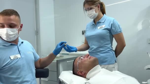 女性看護師は新しい技術で歯を磨くための特別な装置を与えます男の医者は彼の患者が待っている嘘を彼の手にスクロールする手の上に置きます — ストック動画