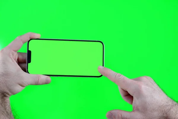 Man holding phone with chroma key display against the green screen background. selective focus Male hands hold a modern black smartphone with green blank screen on neon green background flat lay