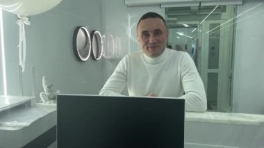 man at the reception desk leans his elbows and looks straight into frame in front of laptop he smiles on him a white sweater handsome adult man 40-45 years old hospital dentistry travel agency