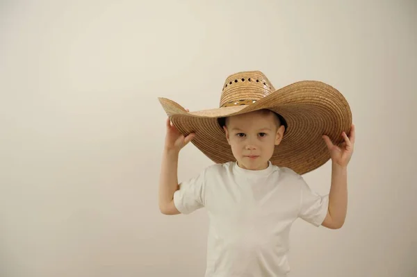 little boy 5 cowboy hat big hat fits over eyes he straightens it t-shirt is isolated in studio white background he smiles spinning makes his eyes beautiful boy place for text clothes headwear