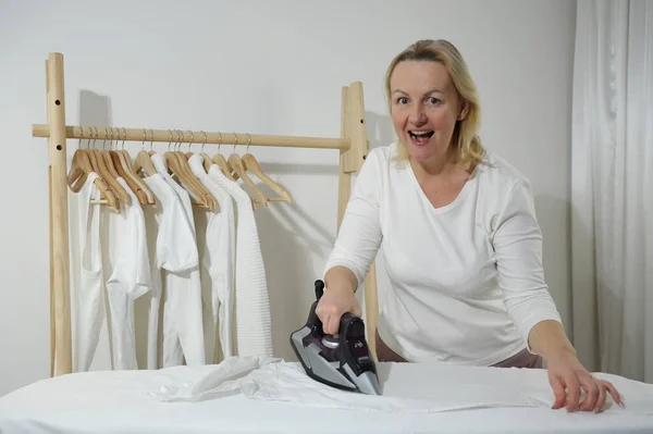 Woman hand ironing clothes isolated woman Smiling with open mouth with shirt ironing while working at home. Silvery dark iron on a white table Background wooden hanger with white clothes