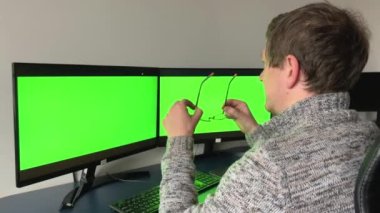 Creative Middle Aged Designer Sitting at His Desk Uses Desktop Computer with Two Green Mock-up Screens. Professional Office Employee Working Late in Evening in Studio Man taking off putting on glasses