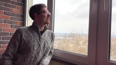middle-aged man in glasses with red beard mustache in gray sweater looks out of high floor window he is calm pensive winter no snow emptiness dampness experience sadness meditation planning brick