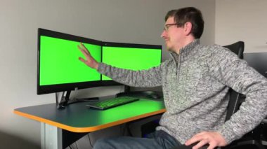 young programmer artist web designer looks at monitor with two green screens chromakey he gently swallows with hand on one of monitors advertisement can be he is very happy after that he laughs hard