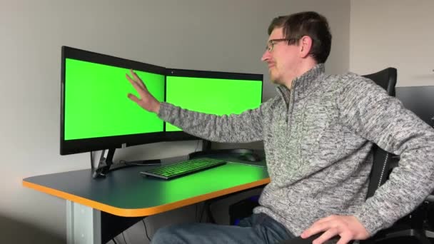 若いプログラマーのアーティストのウェブデザイナーは2つの緑の画面でモニターを見ます彼は優しくモニター広告の一つに手で飲みます彼は彼がハード笑い後に非常に満足していることができます — ストック動画