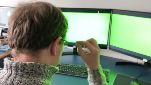 男は2台のコンピュータでオフィスで働いています緑の画面クロマキー彼は後ろに座って 画面を見てメガネを脱いで戻るグレーの部屋赤い髪のプログラマーデザイナー3Dアニメータオフィスの仕事 — ストック動画