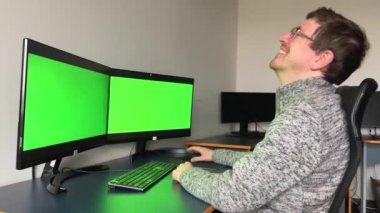 middle-aged man in gray sweater glasses laughs very sincerely strongly he points finger throws head up sits Near table on chair in front two screens 2 monitors with green chromakey screen text ad