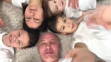 cheerful joyful family lies together on carpet face up shooting from above they wave their hands smiling three boys brothers mom and dad father man bald woman has black hair all dressed White t-shirts