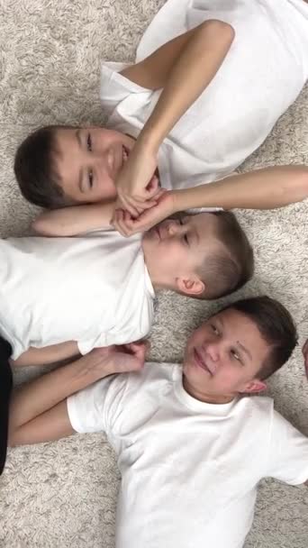 3人の異なる年齢の兄弟は白いTシャツの中にあります白いシャツカーペットの上で遊ぶ笑顔彼らの顔と頭は見える兄は真剣に見える若い1つの演劇真ん中1笑 — ストック動画
