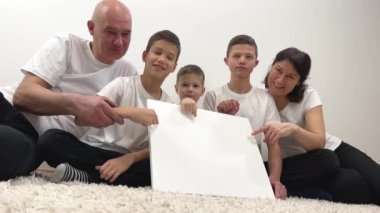 cheerful family large family sits on floor in front of them white square background on which adcan be everyone points finger at especially little boy sits in middle And cutely pokes finger at place