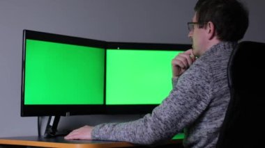 Middle aged with glasses red beard handsome specialist working on desktop computer with green screen mockup in busy creative office with colleagues. Male manager with trimmed beard wears gray sweater