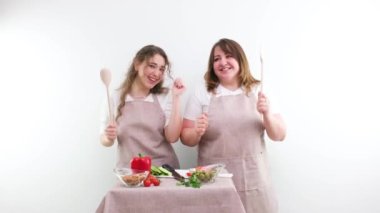 2 women dancing while preparing tasty and healthy food vegetarian food vegetable salad in hands wooden spoons identical kitchen aprons white background joy fun family Cozy weekend mom and daughter