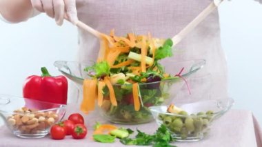 delicious fresh vegetable salad scatters woman stirring in plate lot of carrots greens croutons Parmesan cucumbers on table olives red bell peppers tomatoes different nuts close-up on white background