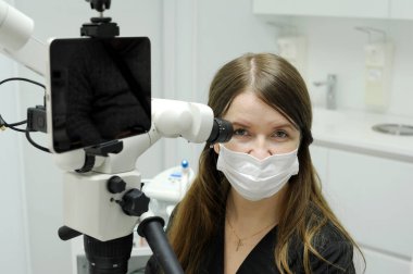 Big screen tablet young dentist sits near microscope for dental treatment research prosthetics filling canals teeth looks into the frame an advertisement for dental clinic on mask latest technology