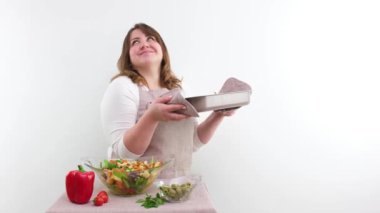 middle-aged woman housewife cook holding tray of food in hands sniffs it smiling showing tasty dance shakes head In foreground vegetables salad ingredients for dinner or lunch at end shows thumbs up