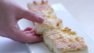 On a square white plate lies a delicious cottage cheese dessert. Homemade cakes woman takes one piece with her hand. Slow motion.