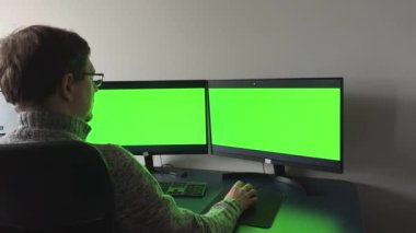 Middle aged with glasses red beard handsome specialist working on desktop computer with green screen mockup in busy creative office with colleagues. Male manager with trimmed beard wears gray sweater