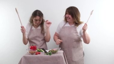 2 women dancing while preparing tasty and healthy food vegetarian food vegetable salad in hands wooden spoons identical kitchen aprons white background joy fun family Cozy weekend mom and daughter