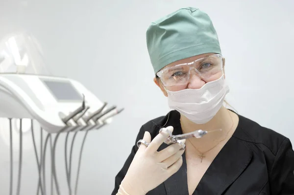 female doctor in a dental office in mask and goggles Green cap to contain hair black suit in her hands carpal syringe and hygienic surgical gloves white wall latest technology cross on the chest