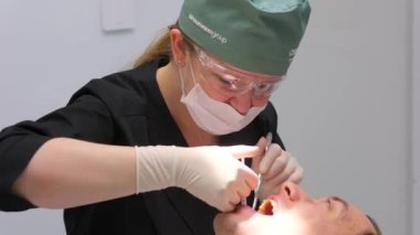 Kadın doktor dişçi karpal şırınga ile anestezi yapar. Sabırlı adam dişlerini aynayla muayene eder ve şırınga verir. Mavi eldivenli asistan hijyenik maske cerrahi eldiven hazırlama