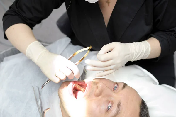 female doctor in black clothes in dentistry sews up wound after a dental operation, a tooth is removed from a man, he looks at her with small eyes, it hurts him anesthesia dental treatment