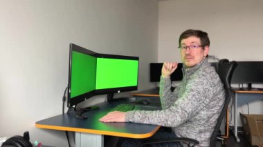Middle aged with glasses red beard handsome specialist working on desktop computer with green screen mockup in busy creative office with colleagues. Male manager with trimmed beard wears gray sweater