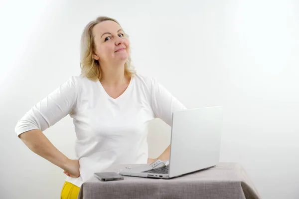 middle-aged woman on white background is standing Near a table with a laptop hands in sideways looks into the frame smiling holding her lips incomprehension surprise expectation space for text