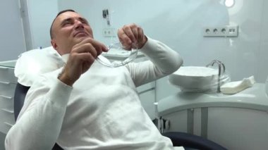 Diş hekimliğindeki adam şeffaf gözlük takar ve beyaz yastığa uzanır. Diş hekiminin protez doldurmasını bekler. Beyazlatıcı beyaz kazak tamir gevşetir.