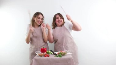 2 women dancing while preparing tasty and healthy food vegetarian food vegetable salad in hands wooden spoons identical kitchen aprons white background joy fun family Cozy weekend mom and daughter