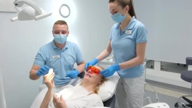 goggles for working with photopolymer lamp are removed by nurse from female patient after procedure in dental office woman is satisfied showing thumbs up smiling gives remote control to doctor