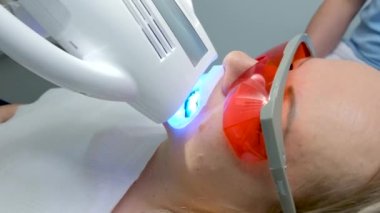Doktor, kadın hasta portakal koruyucu gözlüklerle ilgili diş beyazlatma prosedürünün tamamlanmasını bekliyor. Başının altında ultraviyole rahat yastığa karşı en son teknoloji tedavisini uyguluyor.