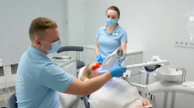 Diş sağlığı kliniğinde dişlerini beyazlatan kadın teçhizat altında yatıyor. Doktor ve hemşire, işlemin tamamlanmasını bekliyor. Elinde panik düğmesi var.