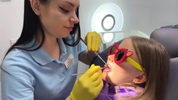 一个无法辨认的儿科牙医的手打电话给坐在医院椅子上的一个笑容可亲的可爱女孩 牙科医生 坐在牙医办公室黄色手套里的小女孩 — 图库视频影像