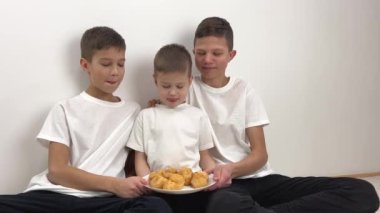 Three cute boys, brothers eat eclairs at home, sunny living room. High-quality shooting in 4k format Three boy brothers sit on the carpet in the room with a plate of eclairs and lick their lips